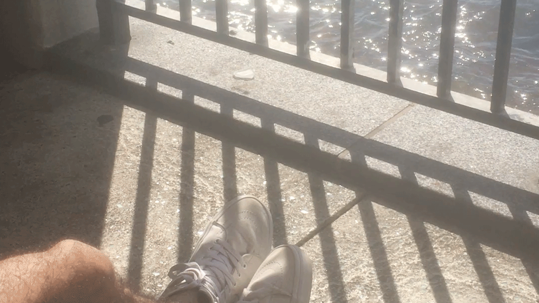 A pair of legs and white canvas sneakers before a railing. Beyond the bars of the railing, water shimmers.