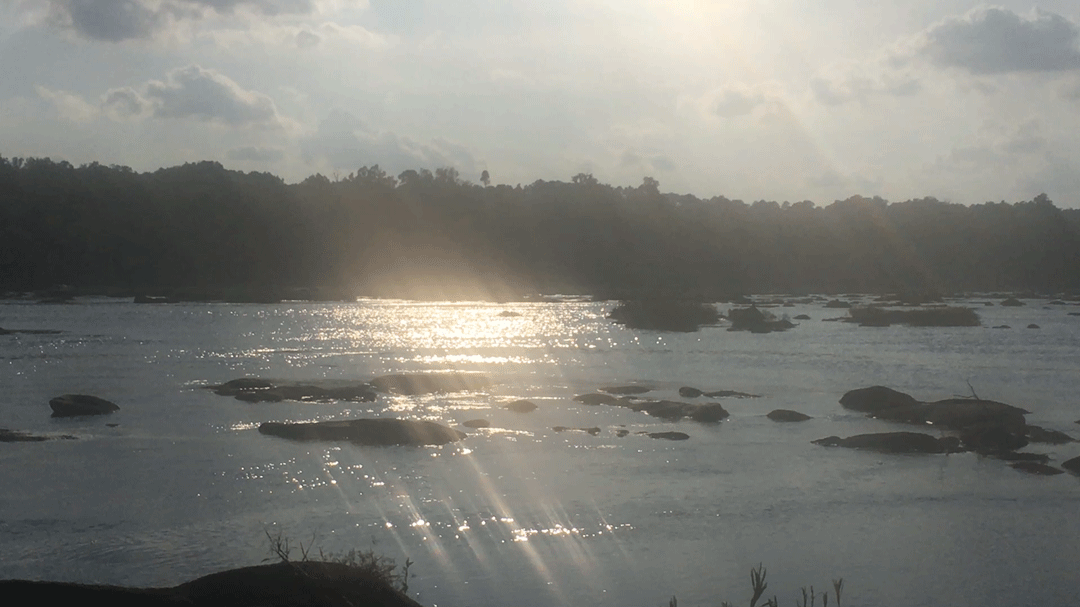 A wide view of a river at dusk, the setting sun streaking across the moving water.