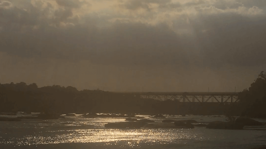 A low river glimmers in the foreground, many large flat rocks laying across it. Cars cross a bridge in the background.