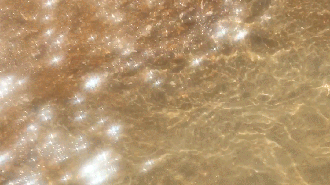 A close-up of waves lapping onto the shore. The sand can be seen through the clear water, and the break is throwing sparkles across the surface.