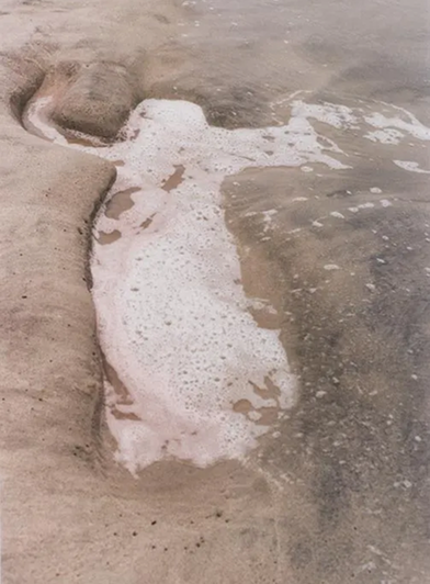 A large indentation in the sand takes the shape of a human body. Tidal waters foam and flow into it.