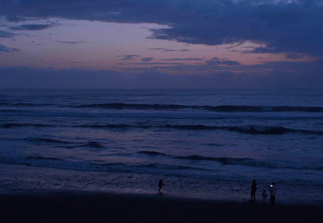 The Pacific Ocean from the shoreline at dusk. A few people stand by water's edge, watching the waves gently lap.