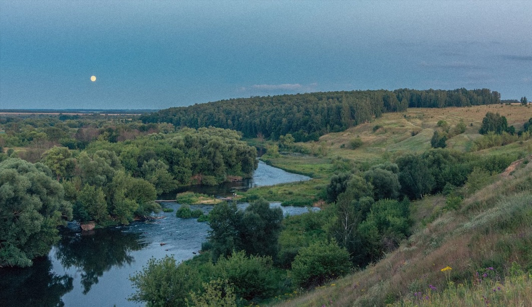 A full moon over a sleepy, tree-lined river, winding toward the horizon.
